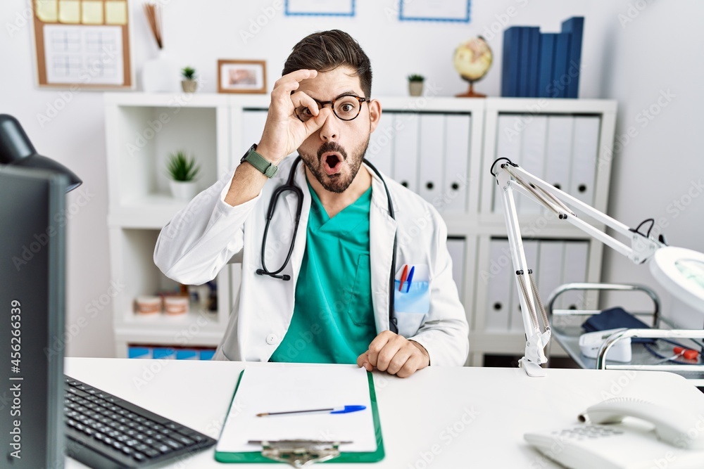 Sticker young man with beard wearing doctor uniform and stethoscope at the clinic doing ok gesture shocked w