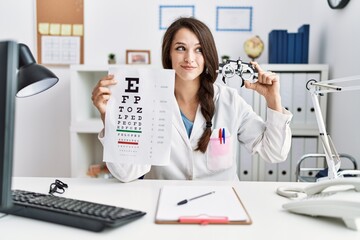 Young doctor woman holding optometry glasses and eyesight test at the clinic smiling looking to the...