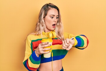 Beautiful young blonde woman drinking cup of coffee wearing headphones looking at the watch time...