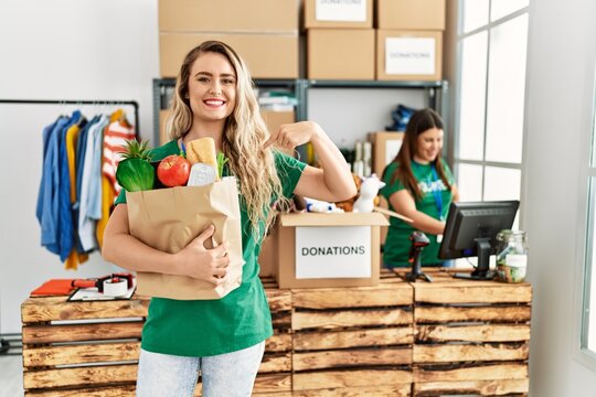 Young blonde woman at volunteer center holding donations paper box pointing finger to one self smiling happy and proud