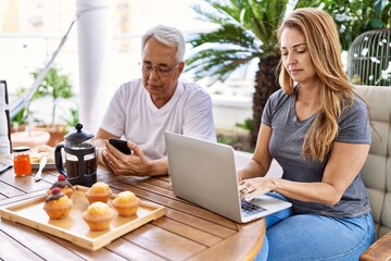 Middle age hispanic couple having breakfast using smartphone and laptop at the terrace.