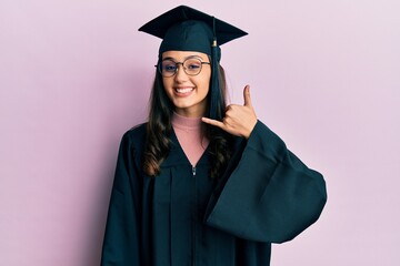 Young hispanic woman wearing graduation cap and ceremony robe smiling doing phone gesture with hand and fingers like talking on the telephone. communicating concepts.