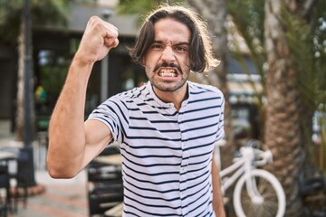 Young hispanic man with beard outdoors at the city angry and mad raising fist frustrated and furious while shouting with anger. rage and aggressive concept.