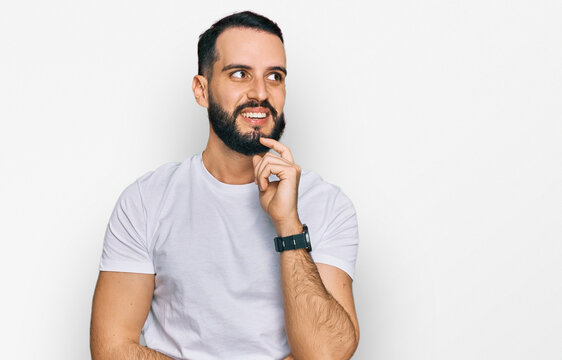 Young man with beard wearing casual white t shirt with hand on chin thinking about question, pensive expression. smiling and thoughtful face. doubt concept.