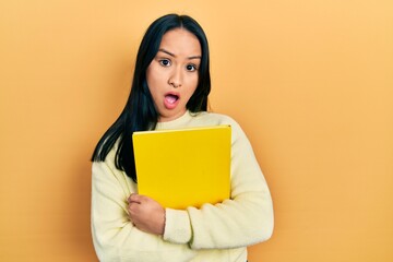 Beautiful hispanic woman with nose piercing holding book in shock face, looking skeptical and sarcastic, surprised with open mouth