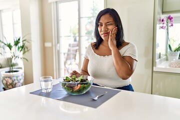 Obraz na płótnie Canvas Young hispanic woman eating healthy salad at home touching mouth with hand with painful expression because of toothache or dental illness on teeth. dentist