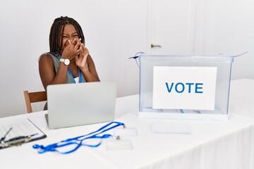 Young african american woman working at political election sitting by ballot smelling something stinky and disgusting, intolerable smell, holding breath with fingers on nose. bad smell