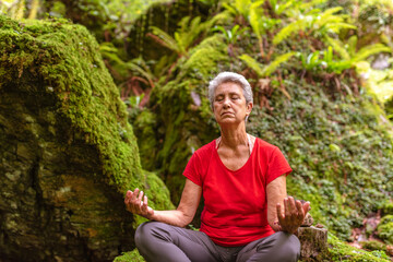 older woman with gray hair and red t-shirt meditating in nature. yoga in the forest