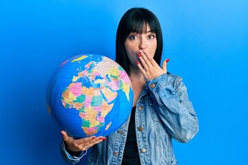 Young hispanic woman holding world ball covering mouth with hand, shocked and afraid for mistake. surprised expression