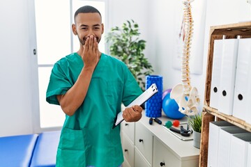 African american physiotherapist man working at pain recovery clinic laughing and embarrassed giggle covering mouth with hands, gossip and scandal concept