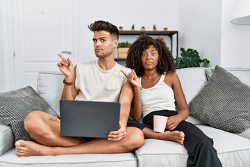 Young interracial couple using laptop at home sitting on the sofa pointing aside worried and nervous with forefinger, concerned and surprised expression