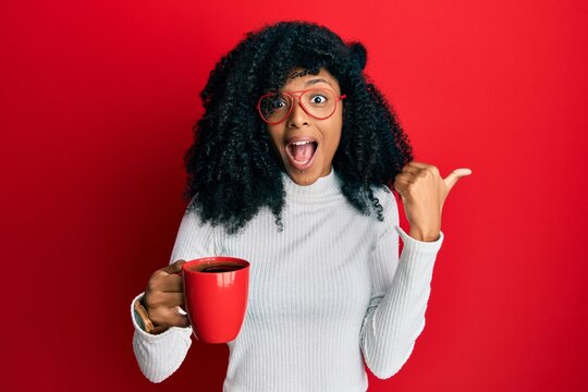 African american woman with afro hair holding coffee pointing thumb up to the side smiling happy with open mouth