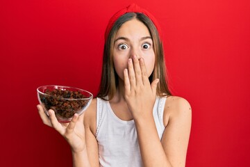 Beautiful brunette little girl holding bowl of raisins covering mouth with hand, shocked and afraid...