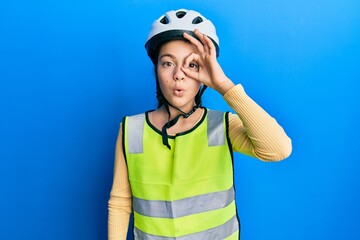 Beautiful brunette little girl wearing bike helmet and reflective vest doing ok gesture shocked with surprised face, eye looking through fingers. unbelieving expression.