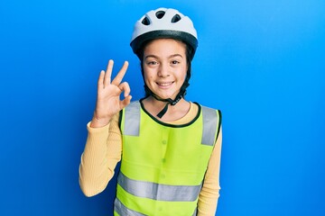 Beautiful brunette little girl wearing bike helmet and reflective vest smiling positive doing ok sign with hand and fingers. successful expression.