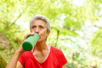 old woman drinking water in the forest. old lady with gray hair doing a mountain route.
