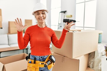 Middle age grey-haired woman wearing hardhat standing at new home showing and pointing up with fingers number eight while smiling confident and happy.