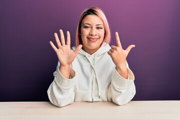 Hispanic woman with pink hair wearing casual sweatshirt sitting on the table showing and pointing...