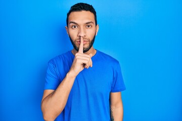 Hispanic man with beard wearing casual blue t shirt asking to be quiet with finger on lips. silence and secret concept.