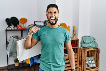 Young handsome man with beard holding shopping bags at retail shop angry and mad screaming frustrated and furious, shouting with anger. rage and aggressive concept.