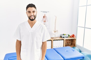 Obraz premium Young handsome man with beard working at pain recovery clinic winking looking at the camera with sexy expression, cheerful and happy face.