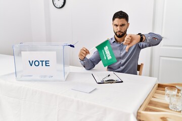 Young handsome man with beard at political campaign election holding arabia saudita flag with angry face, negative sign showing dislike with thumbs down, rejection concept