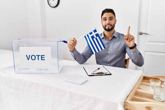 Young handsome man with beard at political campaign election holding greece flag surprised with an idea or question pointing finger with happy face, number one