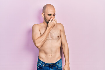 Young bald man standing shirtless smelling something stinky and disgusting, intolerable smell, holding breath with fingers on nose. bad smell