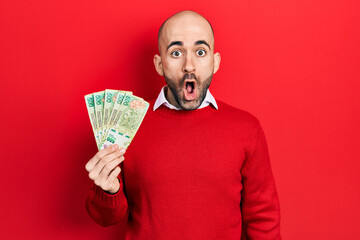 Young bald man holding argentine pesos banknotes scared and amazed with open mouth for surprise,...
