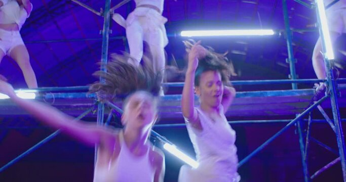 Many female dancers dressed in white shorts dancing inside abandoned building in high scaffolding construction . Stylish Girls dancing, twerking , doing gymnastic movements , jumping . Slow motion 
