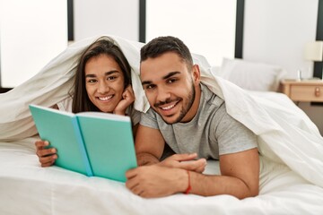 Young latin couple reading book covering with bed sheet on the bed.