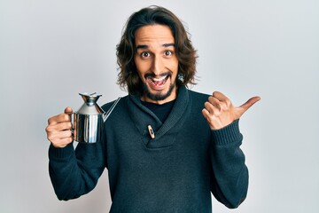 Young hispanic man holding olive oil can pointing thumb up to the side smiling happy with open mouth