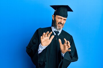 Middle age hispanic man wearing graduation cap and ceremony robe disgusted expression, displeased and fearful doing disgust face because aversion reaction. with hands raised