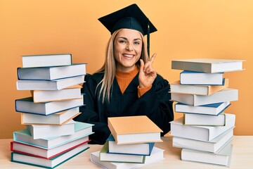 Young caucasian woman wearing graduation ceremony robe sitting on the table with a big smile on face, pointing with hand finger to the side looking at the camera.