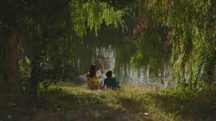 Young and Beautiful couple sitting under the tree by the lake  inside park or forest . Boyfriend and girlfriend throwing stones into water and having fun . Back rear view .  Slow Motion