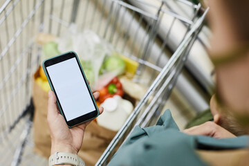 Over shoulder view of unrecognizable woman standing at shopping cart and using mobile app while...