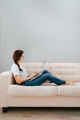 Woman sitting on a bright sofa works and studies at a laptop computer at home.