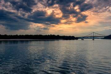 A wide river and a bridge under the setting sun behind the clouds. A calm river in the evening