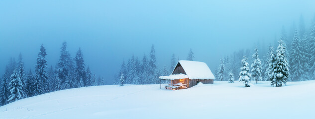 Fantastic winter landscape panorama with glowing wooden cabin in snowy forest. Cozy house in...