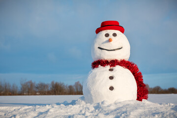 Funny snowman in stylish red hat and red scalf on snowy field. Blue sky on background