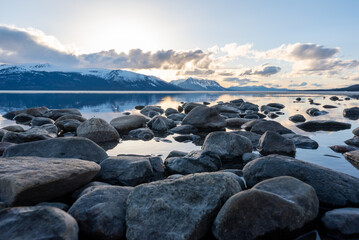 Stunning spring time lake in British Columbia with a lakeshore scene. Ice in distance, rocks and cloudy, blue sky reflection in pristine water. Snow capped mountain peaks in the background. 