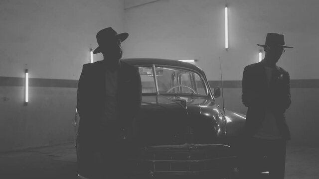 Two silhouettes of men in black suits with classic hats standing by vintage old car. Strangers look like italian mafioso from movie . Footage have permissible count of normally film effect style GRAIN