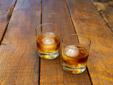 Alcoholic drinks in a clear glass with an ice ball or sphere on a rustic barn wood background.