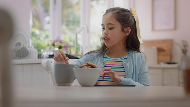 happy little girl using tablet computer eating cookie dipping biscuit into hot chocolate enjoying browsing online relaxing at home