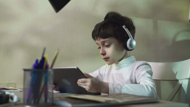 Little Boy Sitting At The Table Listening To Music And Playing Games On The Digital Tablet Watching Video Surfing Internet