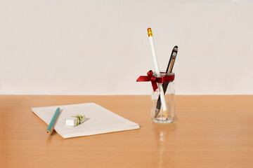Picture of desk with white task pad notebook, pencil, eraser and beside is glass with red ribbon and two pens inside.