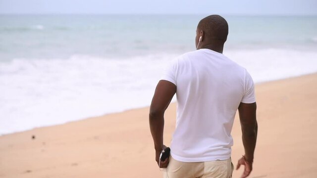 Afro American sporty guy walks along the beach and looks at the sea and listening to music