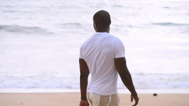 Afro American sporty guy walks along the beach and looks at the sea and listening to music