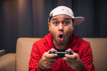 Excited and shocked face of Asian gamer holding headset playing video game online sitting on sofa at living room. Indian professional gamer streaming on social playing game very fun © Chanakon