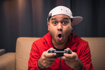 Excited and shocked face of Asian gamer holding headset playing video game online sitting on sofa...